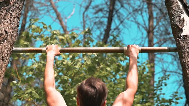 A man performs pull-ups on a horizontal bar positioned between two trees. He shows determination and strength while exercising outdoors under a clear sky and surrounded by green trees