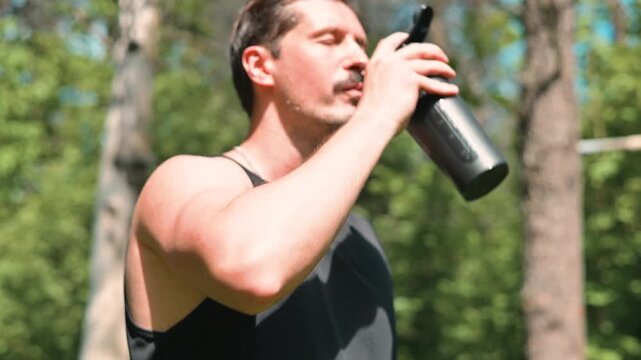 A man in his early thirties, wearing a tank top, is jogging in a forest setting. He takes a moment to drink water and check his fitness tracker during his workout routine