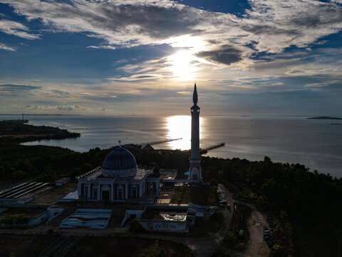 Aerial view of a majestic mosque with a tall minaret and large dome situated on the coastal waterfront during a dramatic sunset over the sea, Kepulauan Riau, Indonesia.
