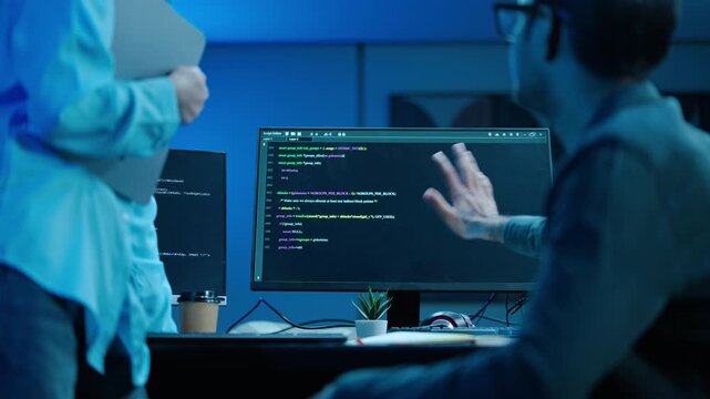 Two professional software developers collaborating on code on a computer monitor in a dark modern office. Male programmer pointing at screen explaining a complex coding issue to his female colleague.