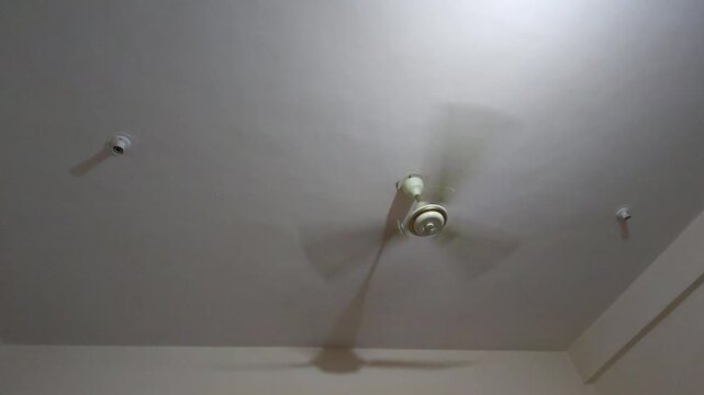 A spinning white ceiling fan casting long shadows above. The nonstop rotation serves as a powerful symbol of escalating global climate crises and the struggle against intense heat waves.
