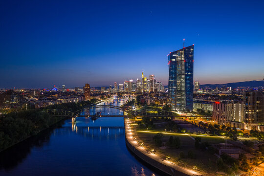 Aerial view of the ECB Tower and the illuminated Frankfurt skyline along the Main river during blue hour with city lights reflecting on the water Frankfurt am Main, Hessen, Germany.