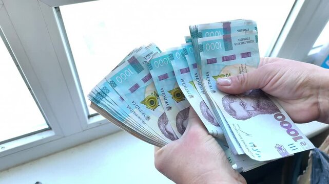 Female hands counting stack of Ukrainian hryvnia banknotes near window. Person holding cash money bill pack closeup. Wealth, financial success, budget planning, currency exchange concept.