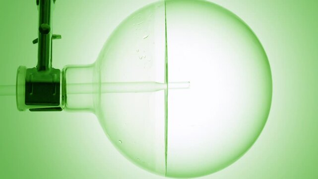 Slow motion close-up of perfectly clear liquid injected through a long dropper into a round-bottom flask, causing swirls and bubbles on green ramp background