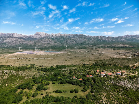 Aerial view of the Suncana elektrana Obrovac solar farm and wind turbines across a rugged landscape with small houses and distant mountains Zaton Obrovacki, Zadar County, Croatia.