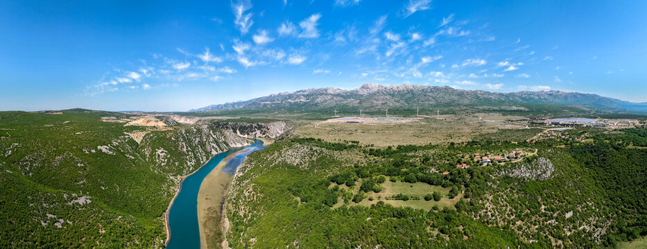 Aerial view of the Zrmanja river canyon winding through green hills with the Suncana elektrana Obrovac and Velebit mountains in Zaton Obrovacki, Zadar County, Croatia.