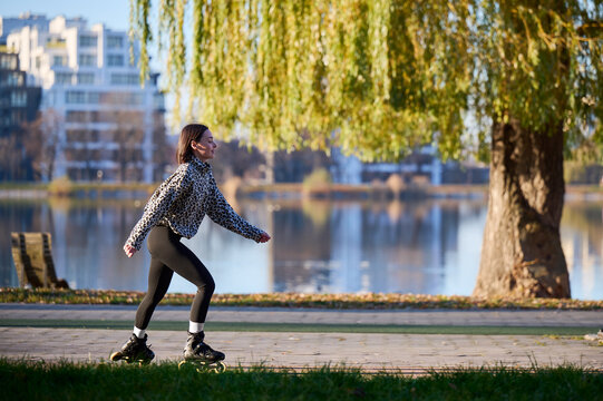 Young female rollerblader enjoying outdoor ride near lake, moving along paved path in soft seasonal light. Suitable for fitness, freedom, recreation or urban wellness promotion