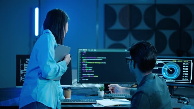 Two diverse tech professionals collaborating in a dark modern office. Asian woman and man discussing programming code and data analytics on multiple computer screens during a late night shift.