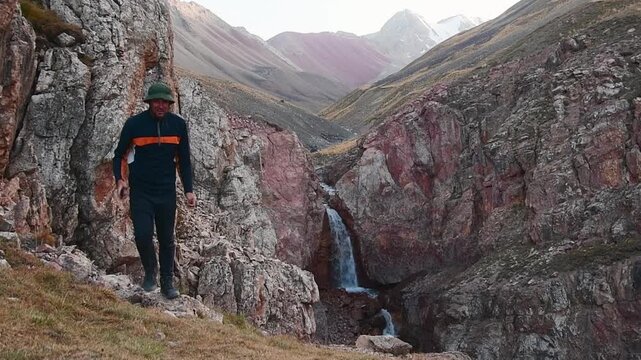 Scenic waterfall in deep rocky gorge near Lenin Peak Base Camp, Kyrgyzstan. Male Hiker viewing taking picture of cascading water in Pamir Mountains. Adventure trekking in Central Asia
