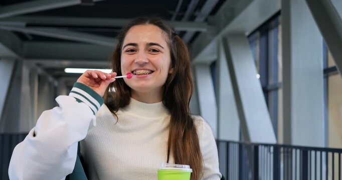 Young white woman eating snack on indoor pedestrian bridge holding green cup wearing white sweater and casual jacket smiling while taking bite of cookie relaxed campus break bright modern architecture