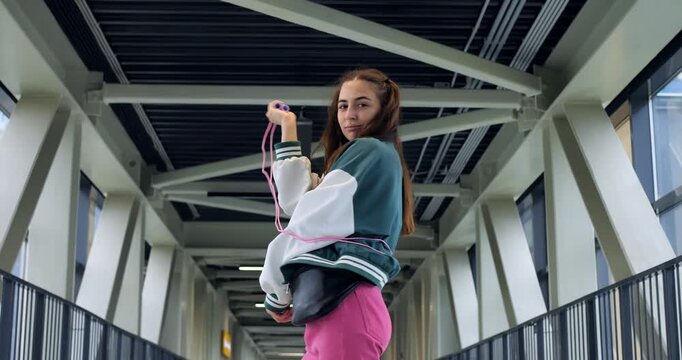 Young woman jumping rope in corridor, energetic workout with pink pants and varsity jacket, rhythmic movement and confident expression, industrial beams and rails frame dynamic fitness scene