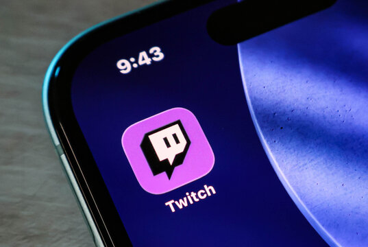 Closeup of the twitch app icon on a smartphone screen, showing the purple logo on a dark background with a digital clock displaying 943