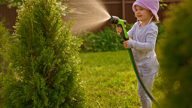Young child wearing a hat and light clothes enjoying watering a green plant with a hose in a sunny backyard garden, helping with chores while learning about plant care