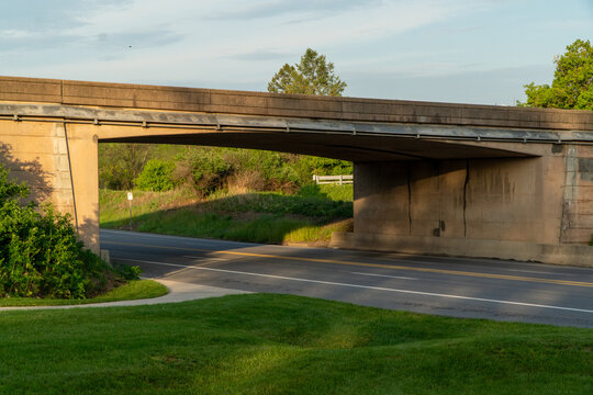 Concrete overpass spanning a quiet road at golden hour, casting long shadows over vibrant green grass and lush trees, with a clear sky above and a small bird in flight.