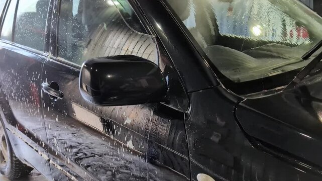 Black vehicle covered in soapy foam receives detailed cleaning. Side mirror, windows, and door panels are visibly coated with suds and moisture.