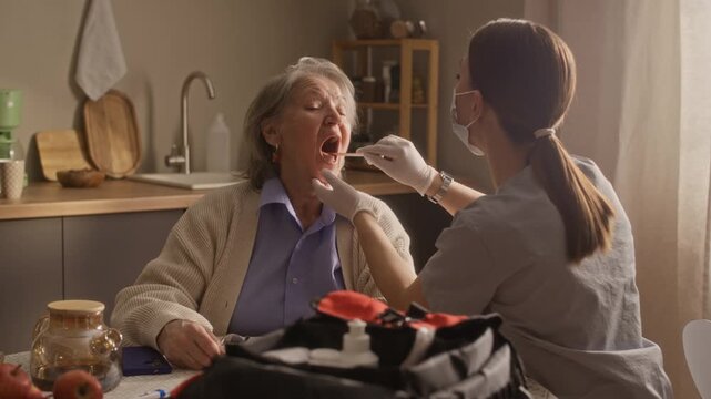 Medium shot of brunette paramedic examining throat of senior woman while they sitting at dining table in kitchen at home
