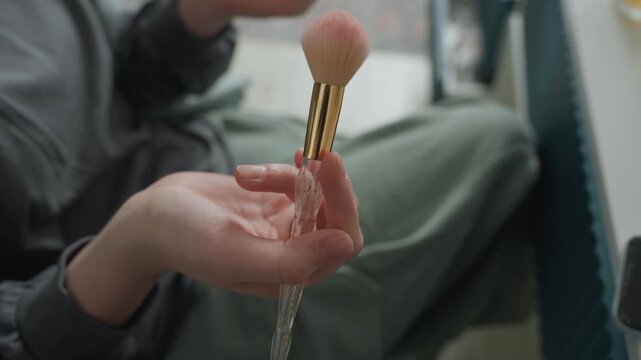 Closeup hand rinsing makeup brush with cascading water droplets pink bristles and golden ferrule catch window light, energetic demo vibe, tactile motion and sparkle, studioadjacent seating, playful