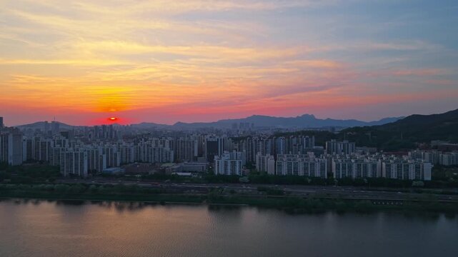 Aerial parallax above Han River bathing in golden sunset light with Gwangjin District and sun setting behind mountains in Seoul South Korea