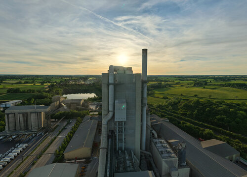 Aerial view of Rugby Cement Plant with its tall industrial chimney and silos against a sunset sky over the surrounding green countryside in Rugby, England, United Kingdom.