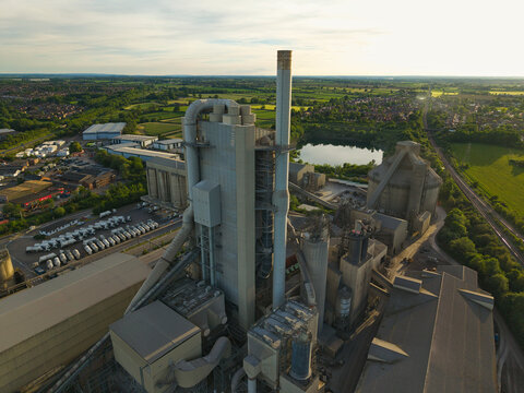 Aerial view of the Rugby Cement Plant featuring tall industrial chimneys and silos surrounded by green fields and a small lake during golden hour in Rugby, England, United Kingdom.