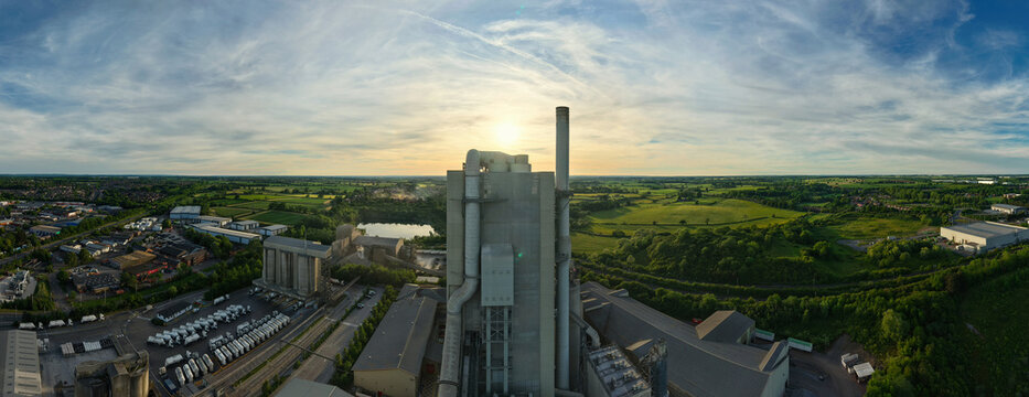 Aerial view of Rugby Cement Plant featuring the tall industrial chimney, silos, and parked trucks under a golden sunset sky in Rugby, England, United Kingdom.