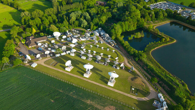 Aerial view of the Satellite Mediaport featuring numerous white parabolic antennas on a green field surrounded by lush trees and a small lake in Long Lawford, England, United Kingdom.