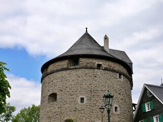 Obraz premium A low-angle medium shot of a massive circular medieval stone tower built from rustic fieldstones under a partly cloudy blue sky. The fortress structure features small defensive windows, arrow slits, a