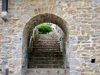 Obraz premium A symmetrical eye-level shot looking through a massive stone archway at an outdoor medieval staircase. The historic stone wall features irregular fieldstones and thick mortar joints, framing the old s