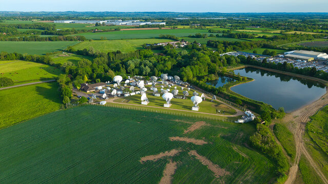 Aerial view of the Satellite Mediaport featuring multiple satellite dishes nestled among lush green fields and a tranquil pond in Long Lawford, England, United Kingdom.