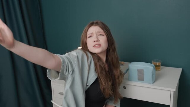 White woman reaching toward camera from desk, pleading expression with outstretched hand, long brown hair, wearing cardigan and crop top, teal wall backdrop, bedside table with jewelry box and lamp,