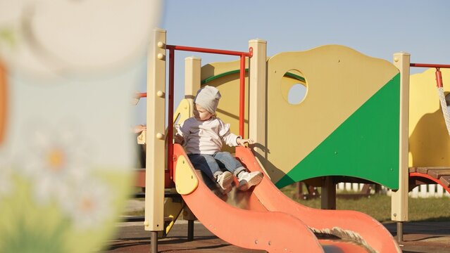 Young girl sliding down an orange and yellow structure, experiencing fun and recreation on a sunny day at a public park, focusing on childhood joy and development