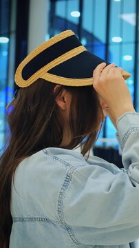 Woman adjusts straw hat in shopping center. Woman experiments with visor accessory near store window display. Female shoppers try different headwear styles while shopping at busy mall kiosk