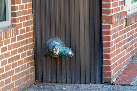 Weathered brass fire department connection standpipe with green verdigris patina mounted on a dark corrugated metal wall, flanked by an old brick building exterior and concrete sidewalk, showing.