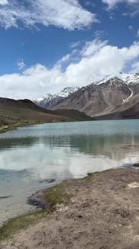 The stunning turquoise waters of Chandratal Lake reflect majestic snow-capped Himalayan peaks under a dramatic blue sky with billowing clouds in the remote Spiti Valley, Himachal Pradesh, India.
