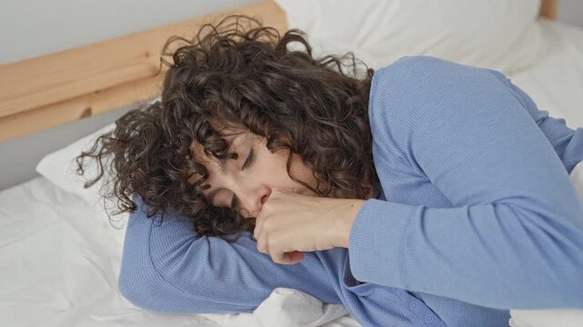 Young hispanic woman with curly hair lying on bed with hand to mouth covering a cough in building; fatigue recovery.