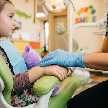 Close up of pediatric dentist hand in blue medical glove comforting and holding hand of little girl patient in green chair children dentistry safety concept