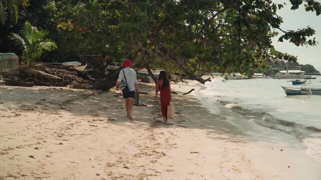 Couple exploring shaded shoreline near boats white man and asian woman walk under canopy of coastal trees carrying straw bag, windblown sand, boat silhouettes, playful discovery and tropical adventure
