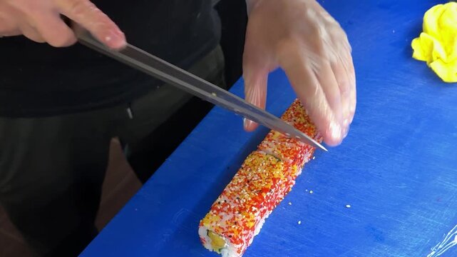 Skilled chef carefully cuts sushi rolls into even pieces using a sharp knife while preparing fresh japanese cuisine on a blue workstation.