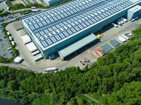 Aerial view of a large industrial warehouse with solar panels on the roof and semi-trucks at loading docks surrounded by lush green trees in Coleshill, England, United Kingdom.