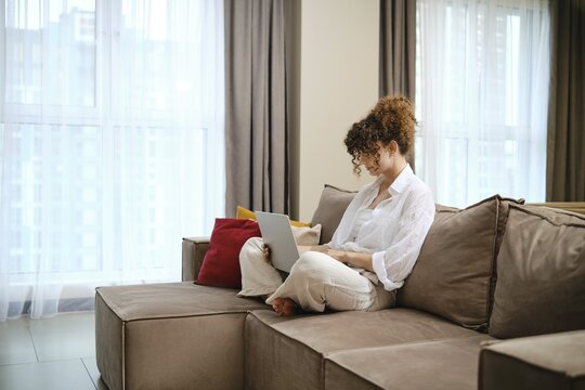 A woman with curly hair is focused on her laptop while sitting on a cozy couch in a bright living room. She makes an online delivery of meal ingredients