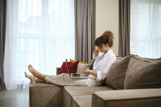 A young woman sits on a comfortable sofa, focused on her laptop, while sunlight streams through large windows