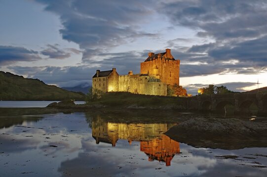 The illuminated Castle Eilean Donan reflected in the water of the Loch, blue hour, film set, Dornie, Highlands, Scotland, Great Britain