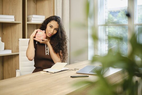In a contemporary office space, a woman with curly hair sits at a wooden desk, smiling as she holds a piggy bank. A notebook and laptop are visible, indicating she is planning her finances