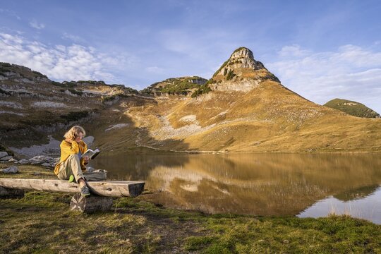 Lake Augstsee and the Atterkogel mountain on the Loser. A hiker sits on a bench and reads a book. Autumn, good weather, blue sky. Altaussee, Bad Aussee, Ausseer Land, Totes Gebirge, Styria, Upper Austria, Austria