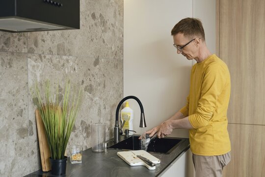 A person in a yellow long-sleeve shirt is washing dishes at a sleek kitchen sink. Sunlight illuminates the modern kitchen with stone and wood elements, creating a warm atmosphere