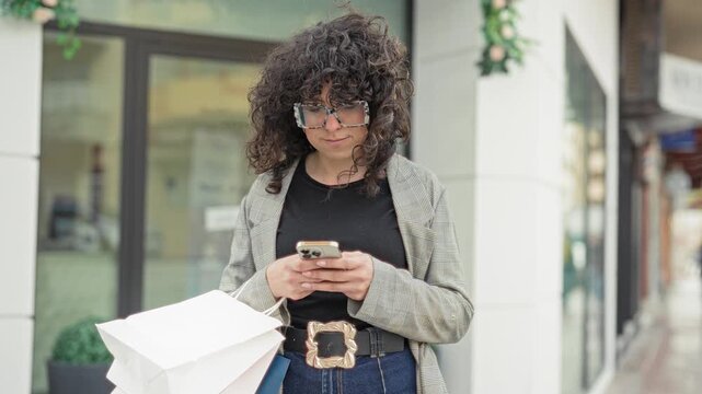 Young woman holding smartphone and shopping bags on street storefront, glancing left while standing near shop window; thoughtful urban shopping mood.