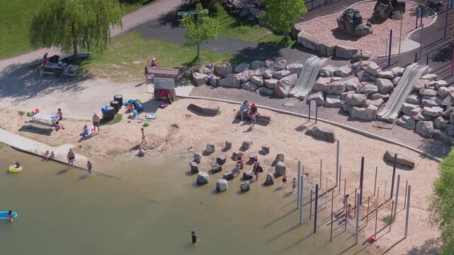 Drone video of people and children playing on the sandy beach at Esther's Pond in Esther Simplot Park, Boise, Idaho. Families enjoying the sunny day with swimming and beach activities.