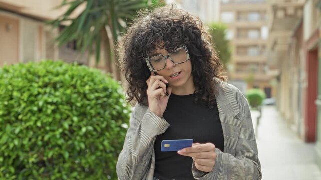 Woman with curly hair and glasses holding smartphone to ear and checking a creditcard with her hand on the card on a street; financial concern.