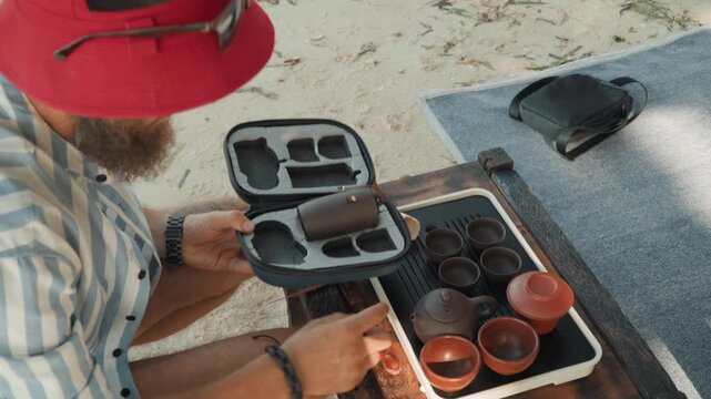Beach tea kit caucasian man unpacking case revealing clay teapots and cups, hands organizing compartments on mat, overhead view, sunlit textures, product lifestyle, careful craft focus