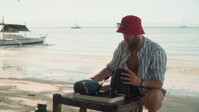 Beach tea ceremony with caucasian man pouring from teapot on wooden table, wearing red bucket hat, boats on horizon, calm waves and warm sunset, solo traveler brewing mindful ritual